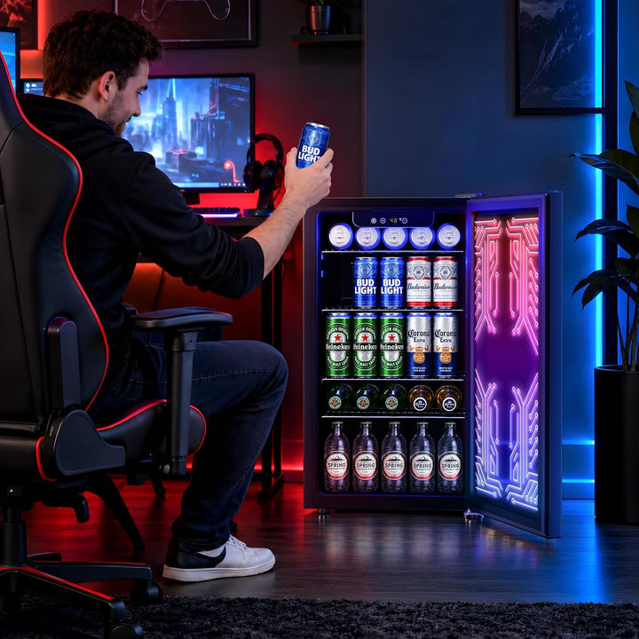 A man sits in a gaming chair holding a can of beer next to an open compact beverage refrigerator filled with canned drinks. The fridge has a glowing purple and pink geometric door panel and shelves stocked with assorted canned beverages.