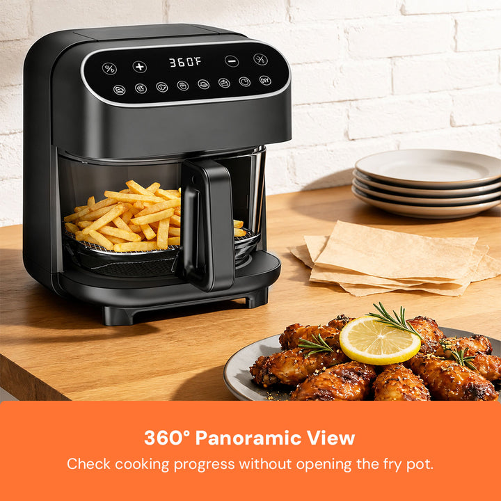 A black countertop air fryer with a transparent window showing french fries inside, placed on a wooden table in a kitchen setting. Plates, napkins, and cooked chicken are nearby, and an orange text banner at the bottom reads '360° Panoramic View Check cooking progress without opening the fry pot.