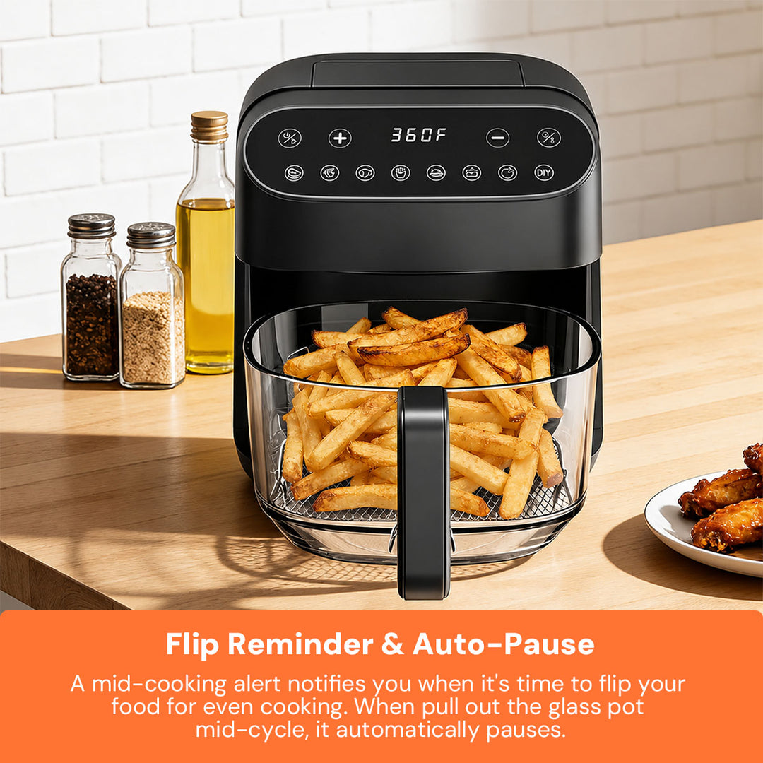 A black air fryer with a glass basket filled with golden french fries sits on a wooden countertop in a kitchen setting. Salt and pepper shakers, a bottle of oil, and a plate of chicken wings are nearby.