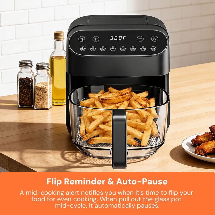 A black air fryer with a glass basket filled with golden french fries sits on a wooden countertop in a kitchen setting. Salt and pepper shakers, a bottle of oil, and a plate of chicken wings are nearby.