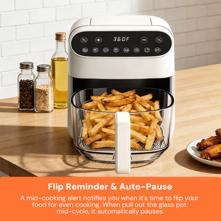 A white air fryer with a glass basket filled with french fries sits on a wooden countertop in a kitchen setting. A digital display on the front shows 360F, and seasoning bottles and a plate of chicken wings are nearby.