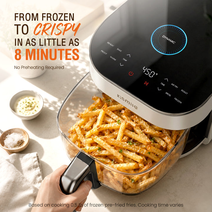A Kismila dynamic air fryer with a clear basket filled with golden crispy french fries, shown with a hand holding the handle. The background is a clean, sunlit kitchen counter with small bowls of seasoning and dipping sauce, accompanied by promotional text.